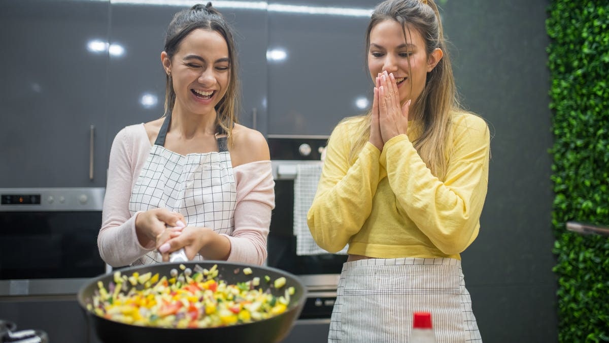 Two women enjoying cooking together in a stylish kitchen, laughing and preparing vegetables in a frying pan.